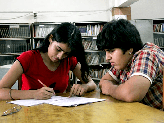 Volunteer scribe in red shirt writing exam answers for student in checkered shirt in library setting with bookshelves in background